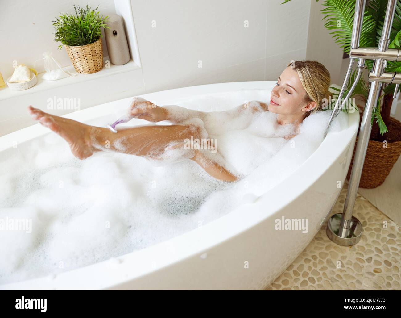 Beautiful woman shaving leg with razor in bathtub with foam. Relaxation