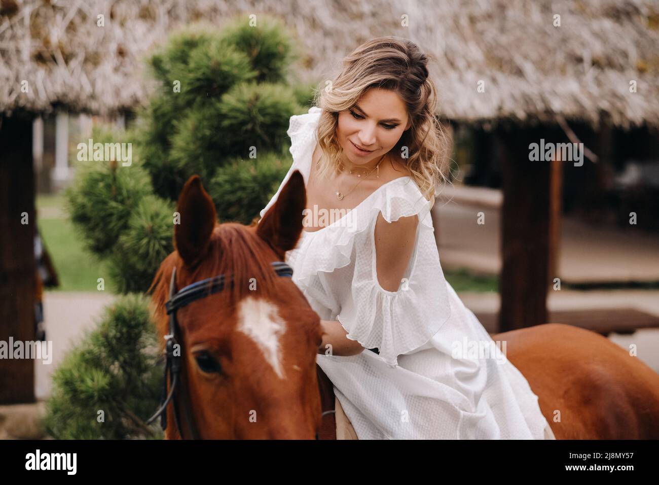 A woman in a white sundress riding a horse near a farm Stock Photo