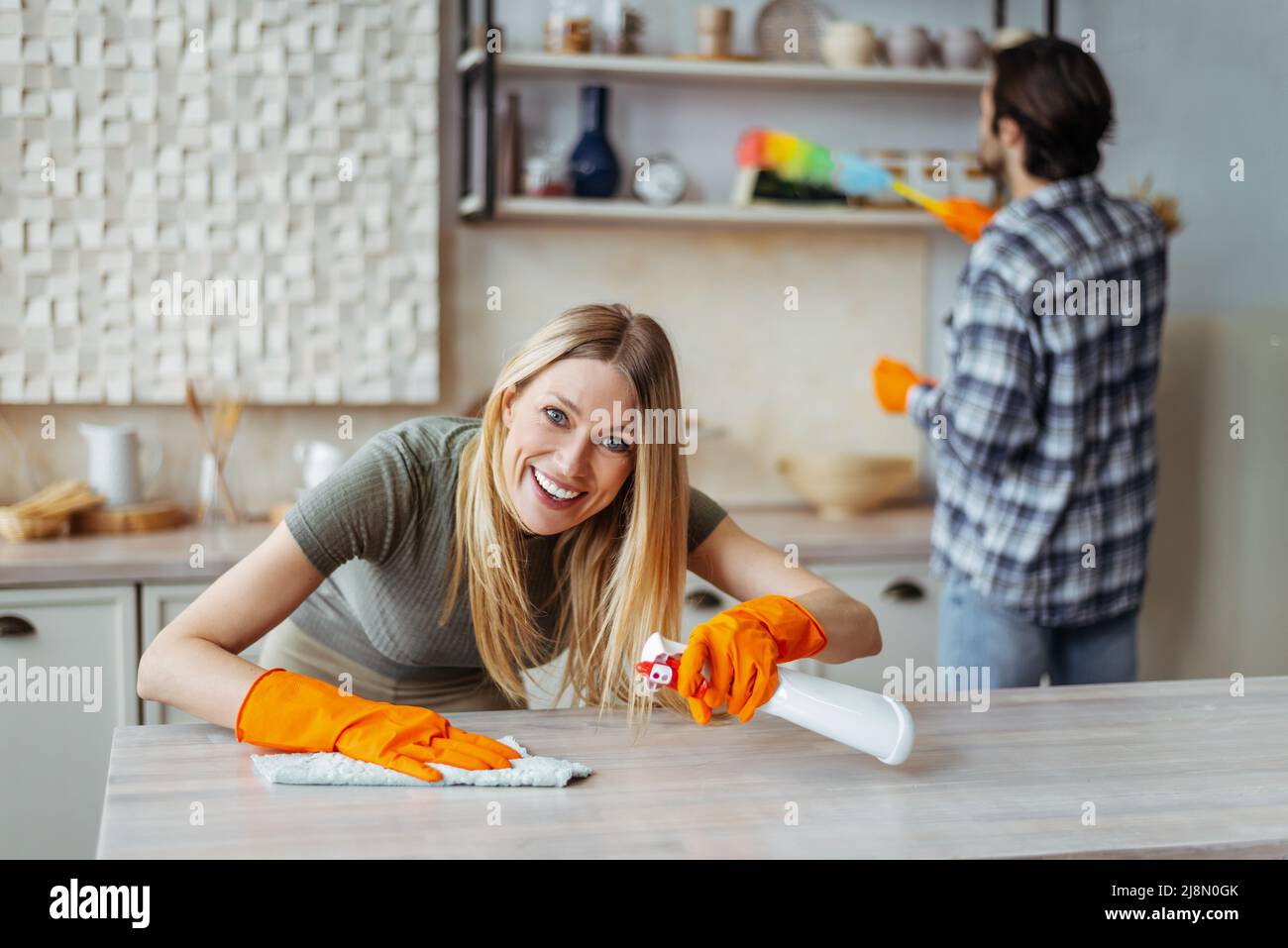 Happy funny young caucasian blonde woman in rubber gloves with spray and husband dust off furniture Stock Photo