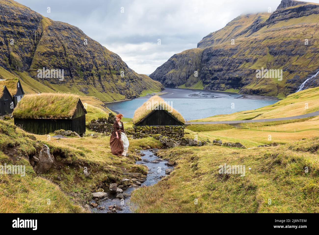 A young redhead woman sits near a stone fence in old-fashioned clothes. Saksun village and sandy shore of the lagoon with an azure lake and dramatic c Stock Photo