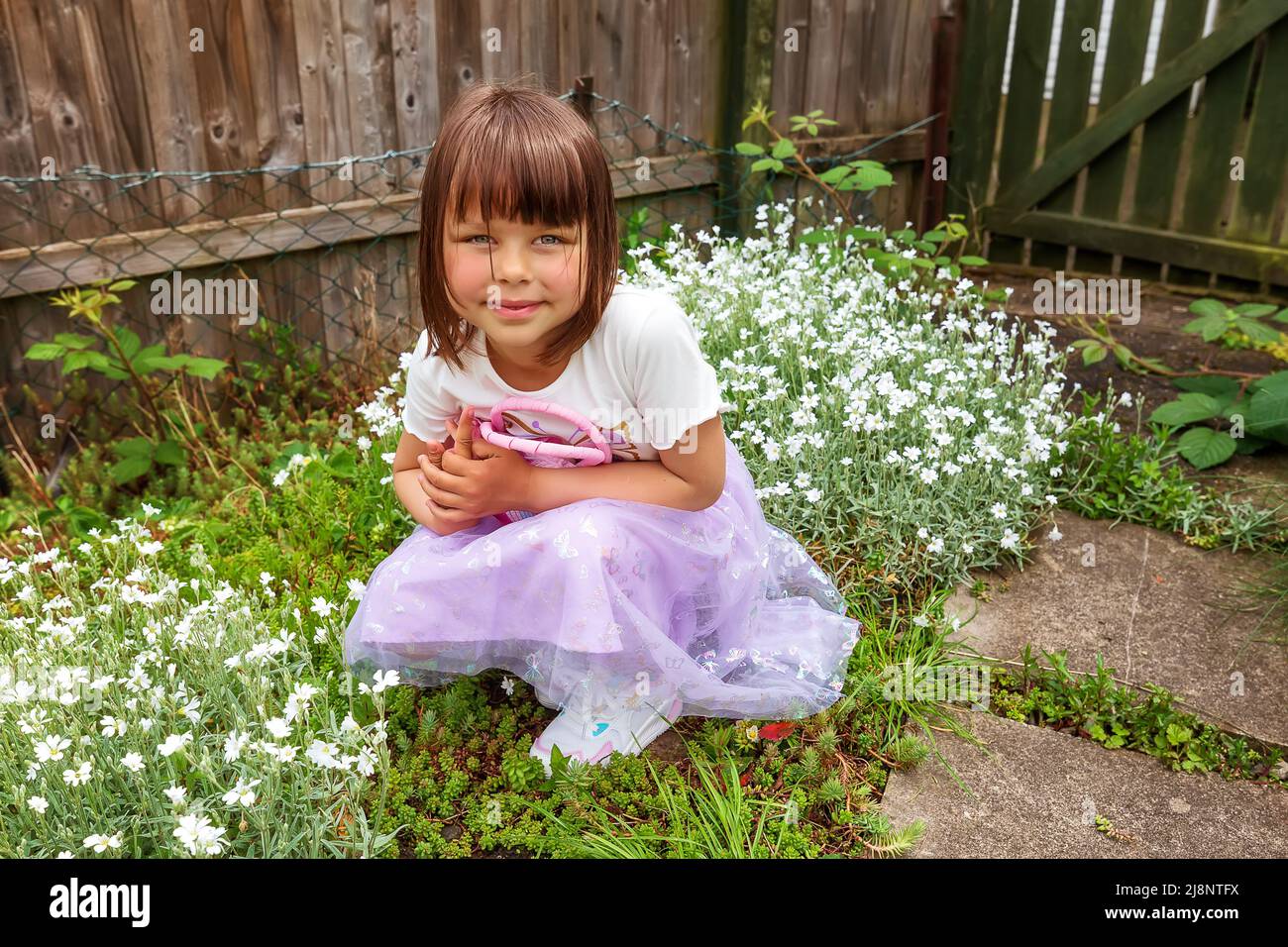 A cute smiling girl is playing in the backyard with small pink bag. Sunny day. Good spring weather Stock Photo