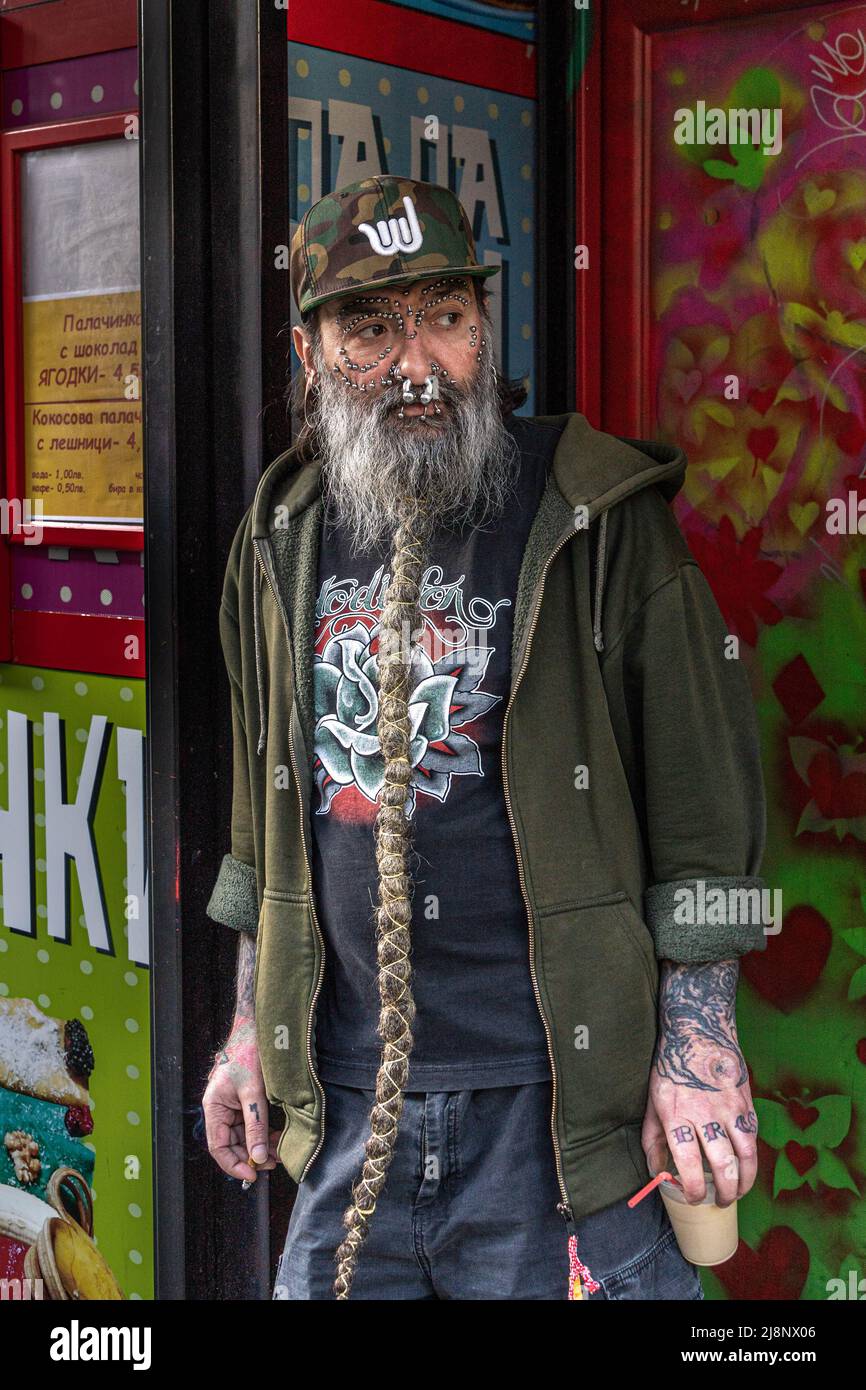 Heavily pierced man with a long grey braided beard, dressed casualy, stands in a doorway with a cup of coffee in his h Stock Photo