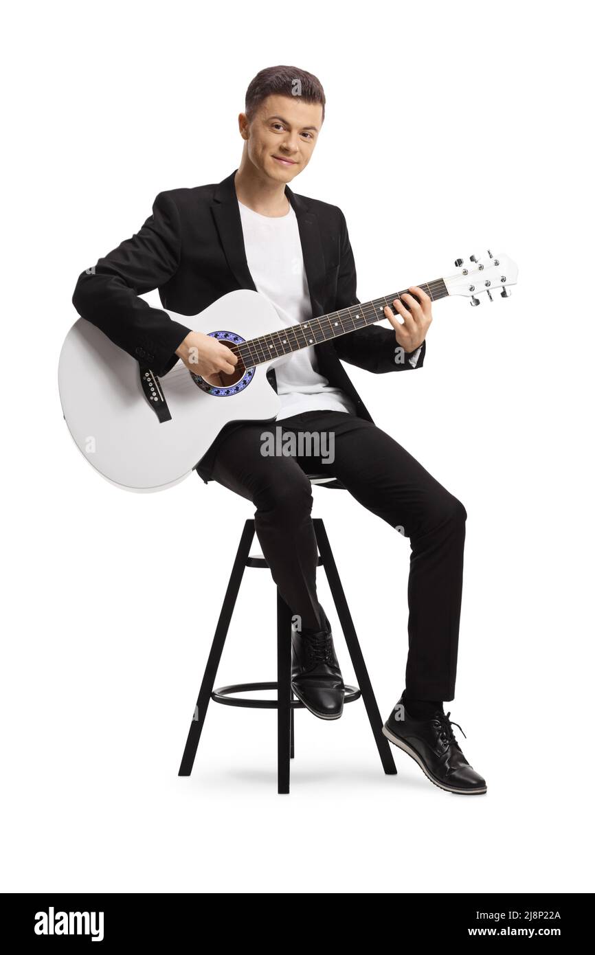 Elegant young man in a black suit playing an acoustic guitar seated on a chair isolated on white background Stock Photo