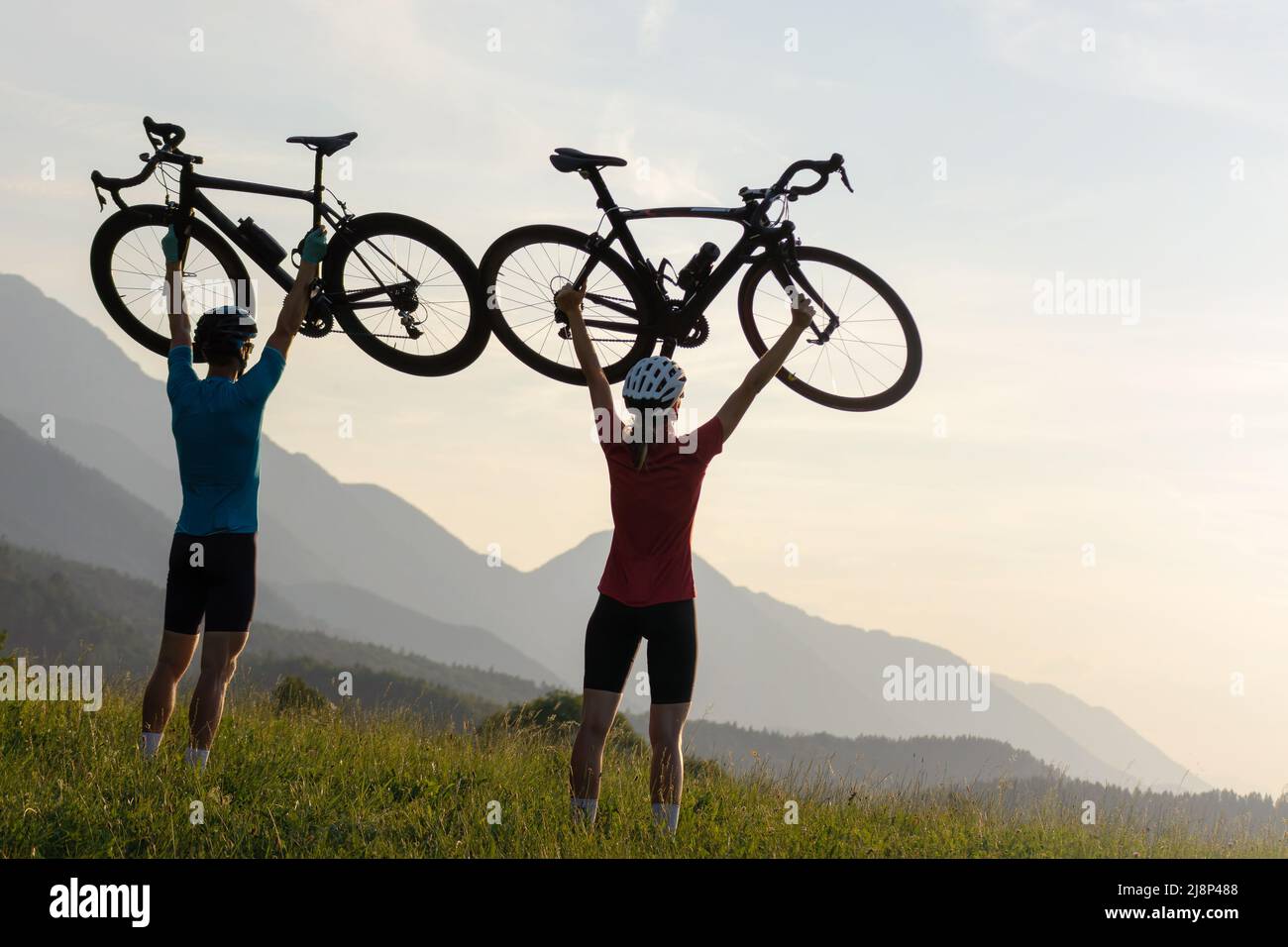 Two cyclers silhouettes holding road bicycles high above heads, in winner pose during sunset over the mountain landscape Stock Photo