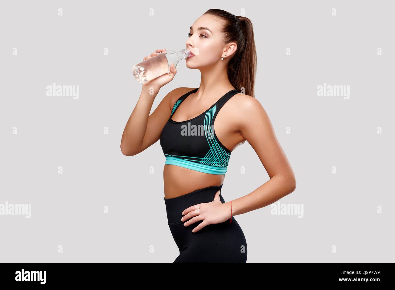 Attractive asian sporty caucasian woman in black fitness suit drinks water on isolated gray background Stock Photo