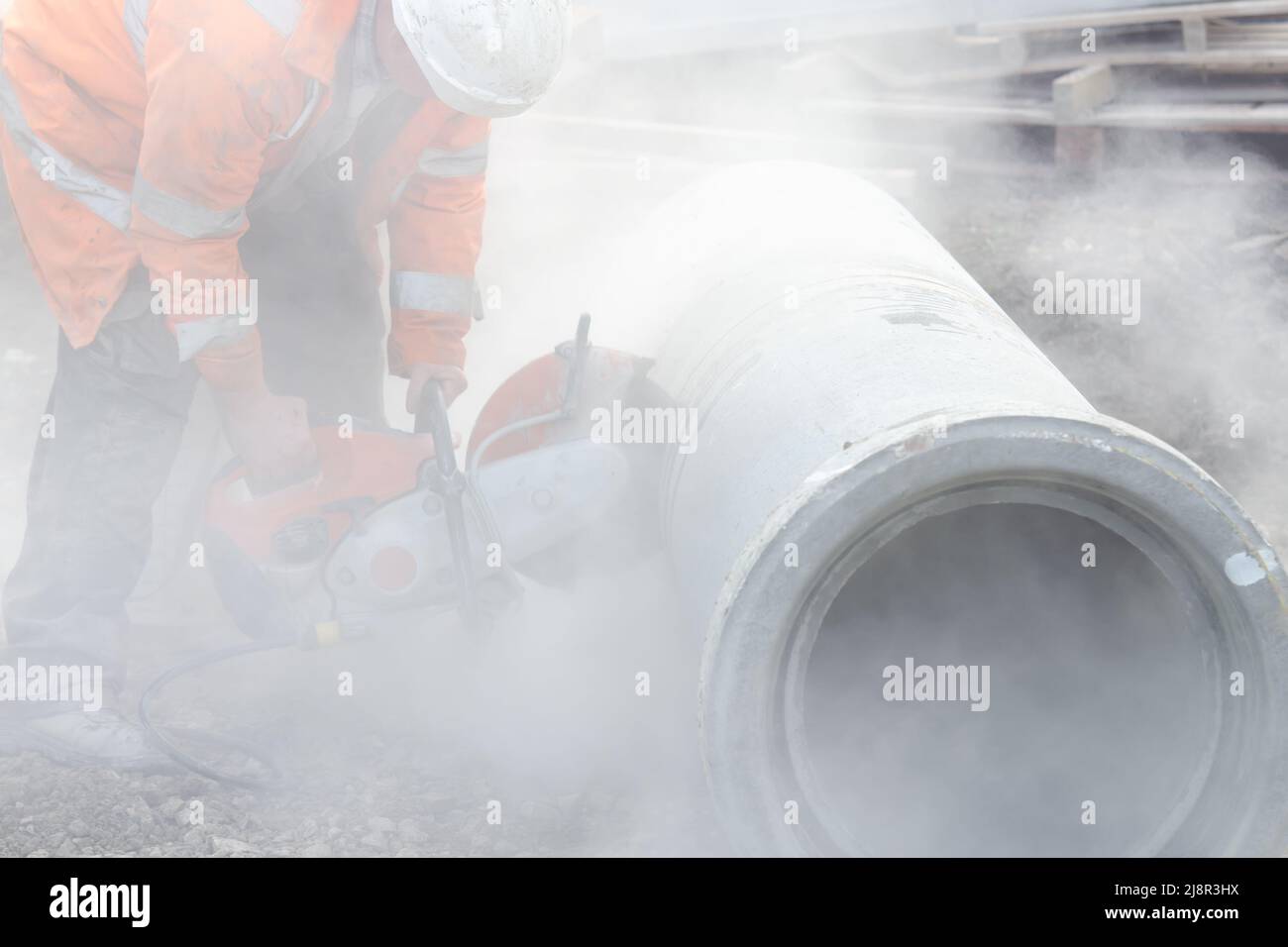 A worker at the construction site cutting a concrete drainage pipe with concrete cutter.Builder covered in a harmful dust cloud as safety procedure br Stock Photo