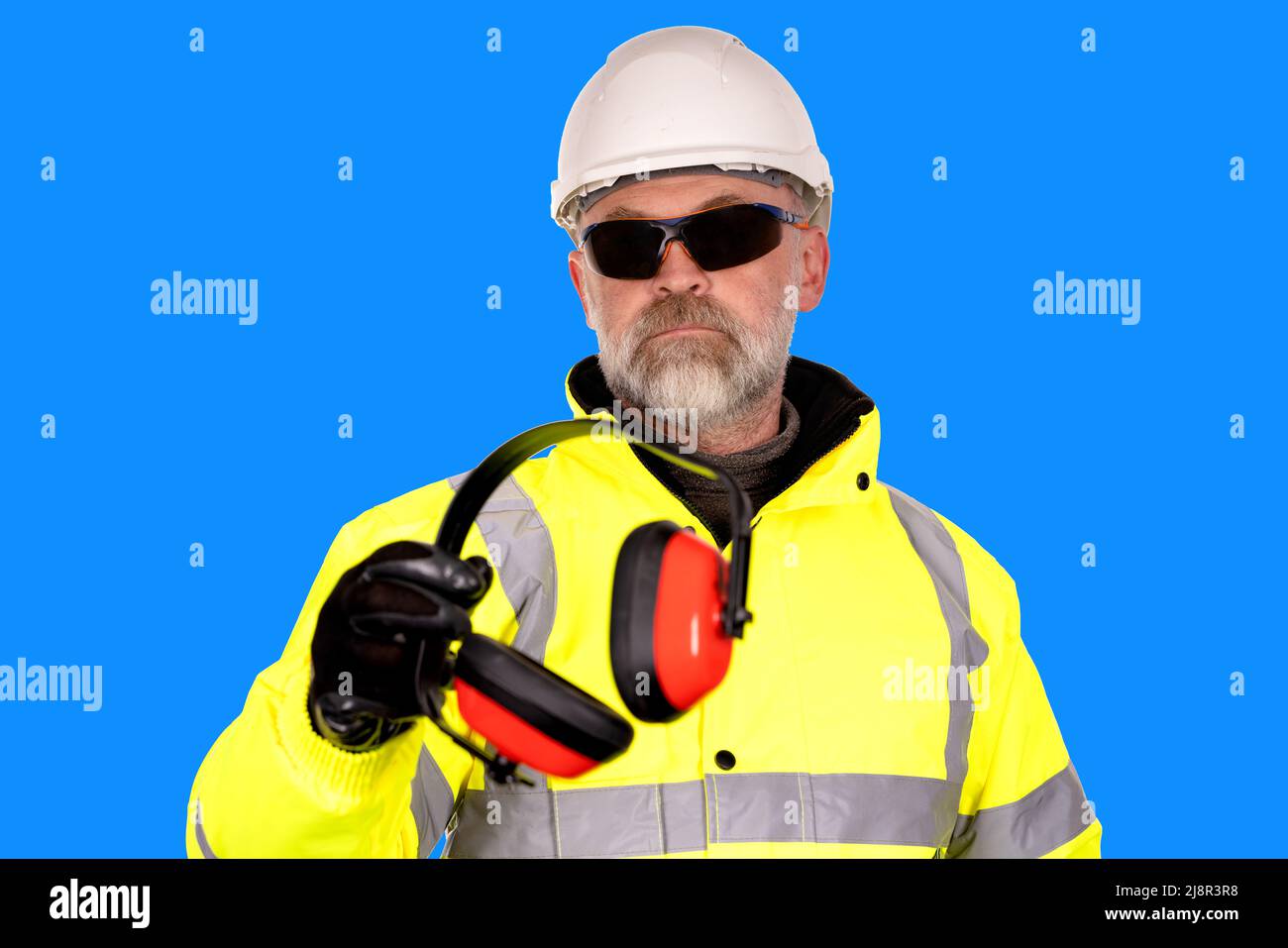 construction worker in hat yellow hi-viz coat and dark safety glasses on blue background gives foam-filled padded ear defenders to viewer Wear hearing Stock Photo