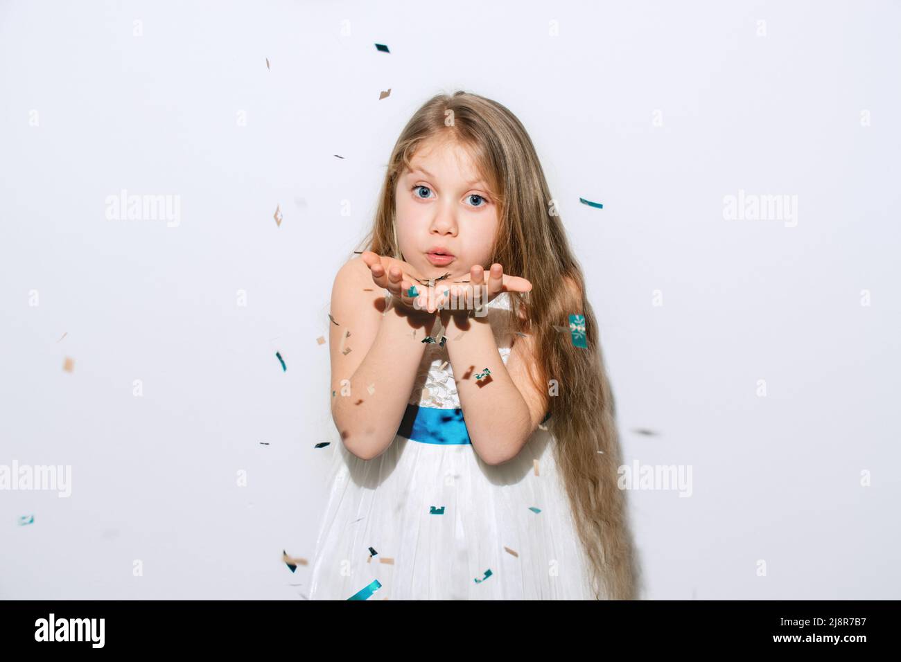 Happy Birthday. Beautiful little girl inflates confetti on a white background. The child is wearing a white dress and a blue ribbon. Big blue eyes and long blond hair. Happy New Year. Copy Space Stock Photo