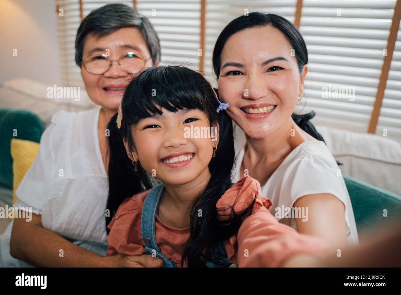 Cute Asian girl smiling and looking at camera while taking selfie with grandmother and aunt while sitting on sofa in living room at home Stock Photo