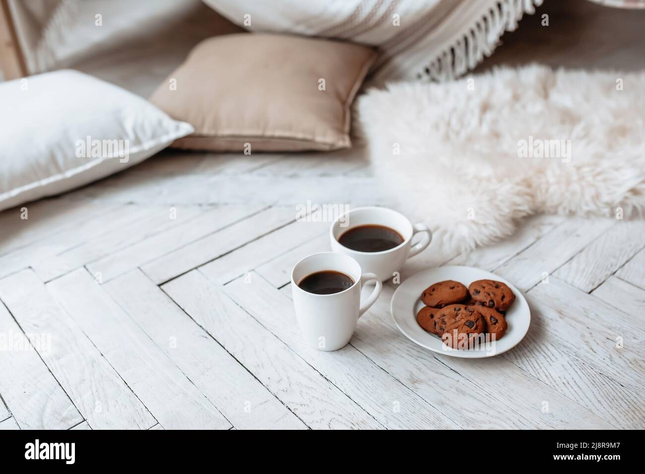 Breakfast in the morning. Two white cups of coffee and chocolate chip cookies stand on a light wooden parquet and pillows lie nearby. Homeliness, day Stock Photo