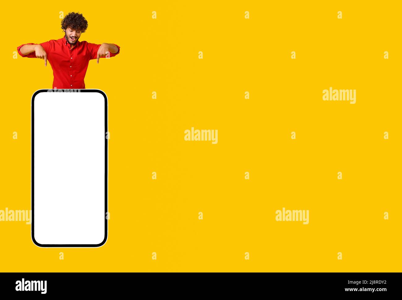 Excited curly Indian young man pops up like gin above smartphone and points finger down at empty screen isolated on yellow background, latin guy presenting and advertising mobile app, copy space Stock Photo