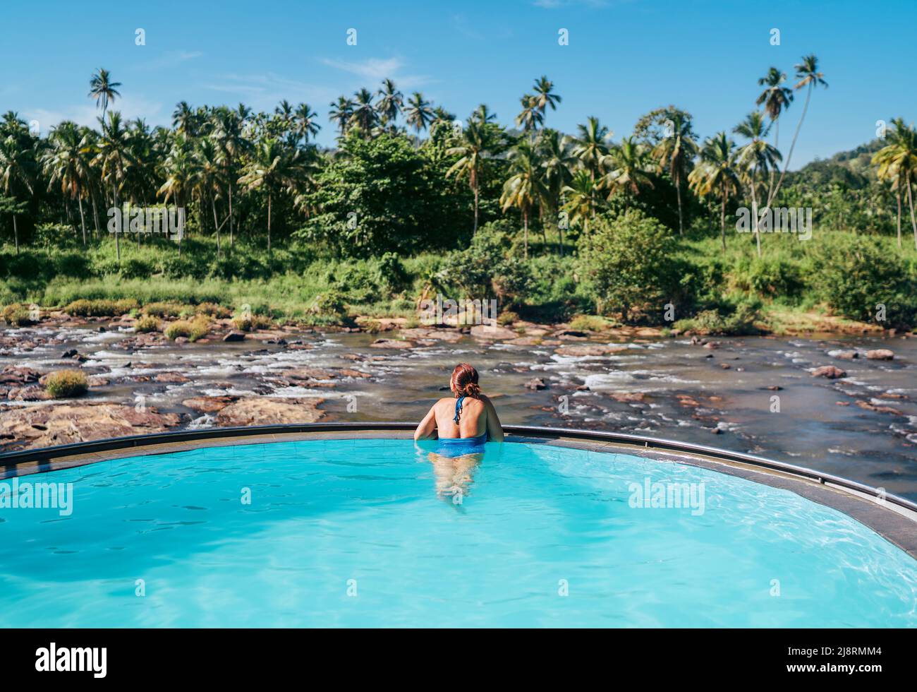 Woman in swimsuit relaxing in infinity swimming pool and watching wide rocky river landscape with jungle banks in Pinnawala Elephant Orphanage. Stock Photo