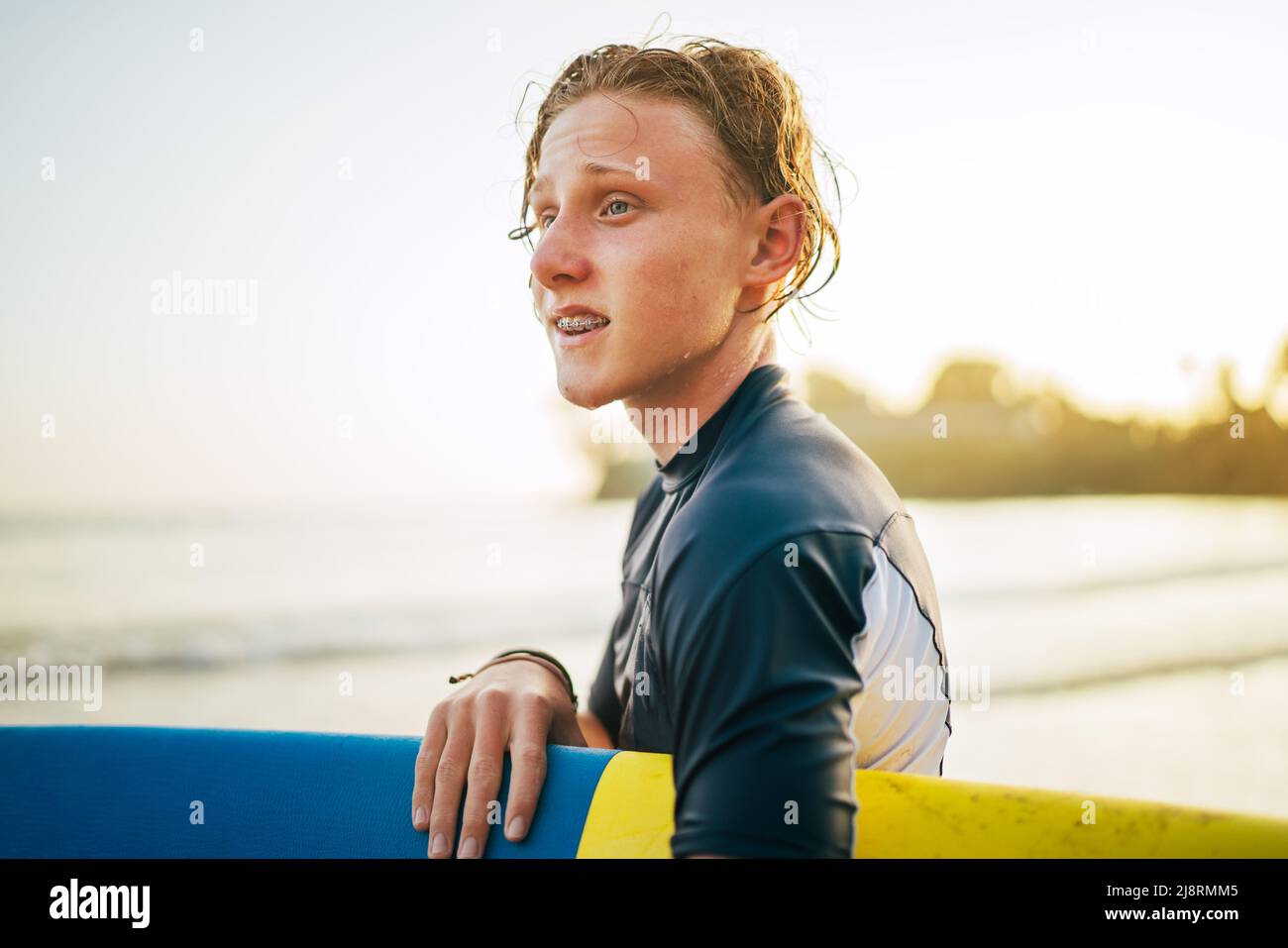Portrait of a teen boy with Dental braces and wet hair with a surfboard goes for surfing. He is smiling and walking into the water. Happy boyhood and Stock Photo