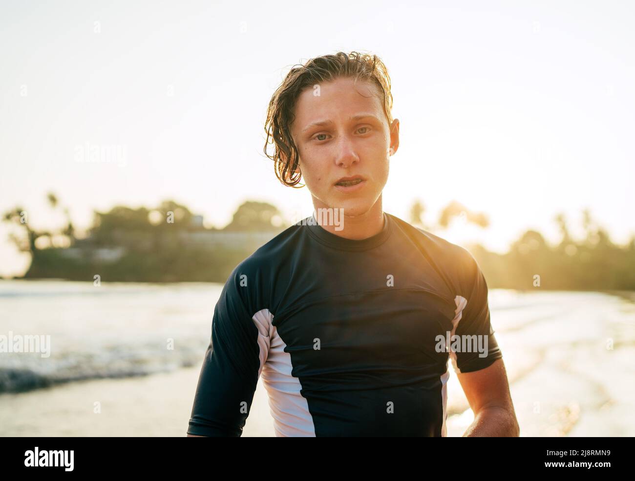 Portrait of a handsome teen boy surfing rashguard on the sand Indian ocean beach with sunset on background. Happy teen time and active vacation time c Stock Photo
