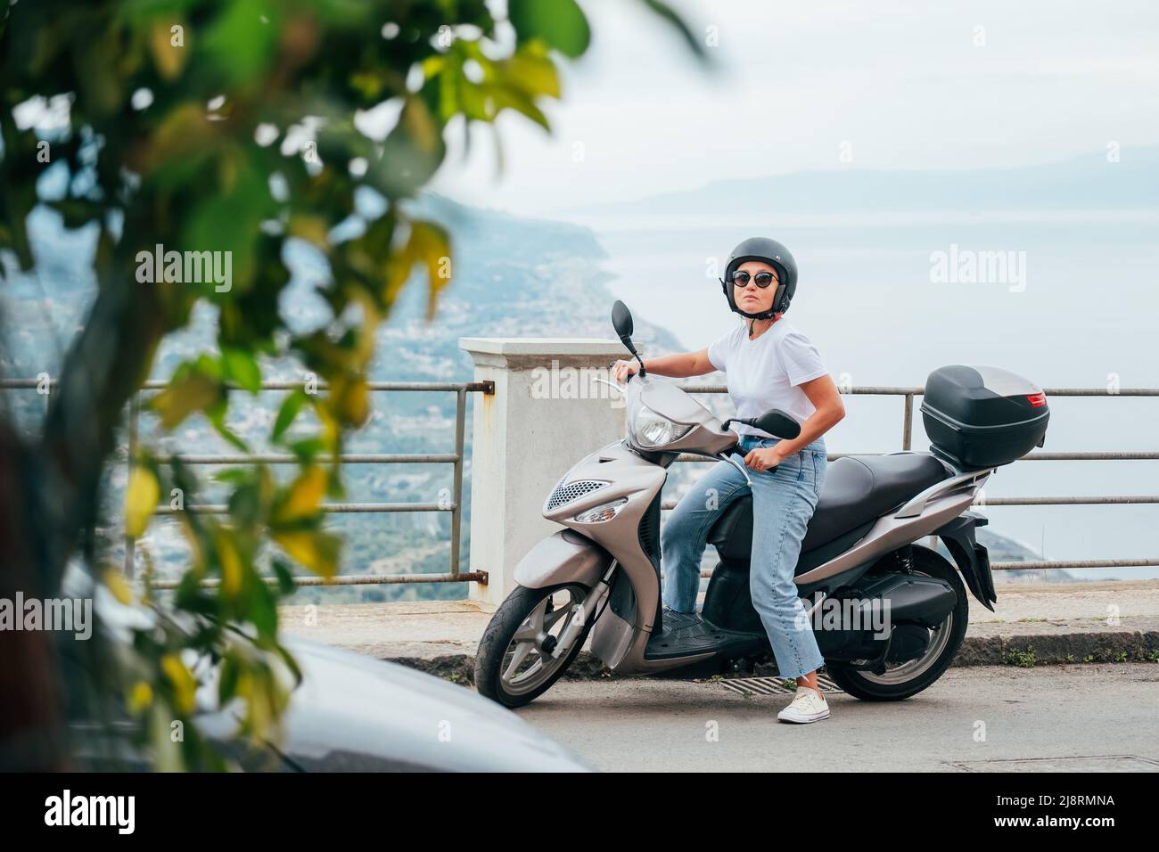 Middle-age woman in helmet and sunglasses on motor scooter on the Sicilian old town streets in the Forza d'Agro with Sant'Alessio Siculo harbor. Happy Stock Photo