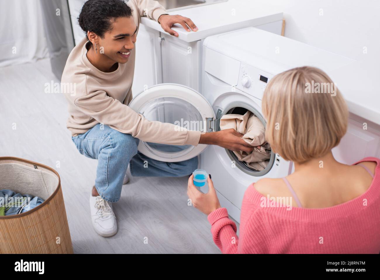 happy blonde woman holding cup with detergent near african american man putting clothes in washing machine Stock Photo