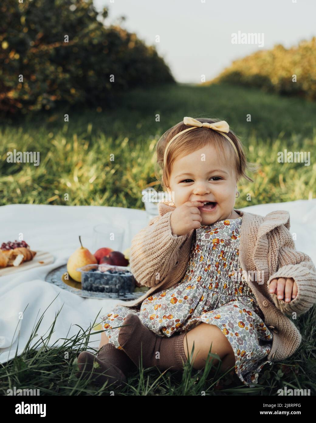 Vertical Portrait of Cheerful Little Baby Girl Sitting on a Plaid on Picnic Outdoors at Sunset Stock Photo