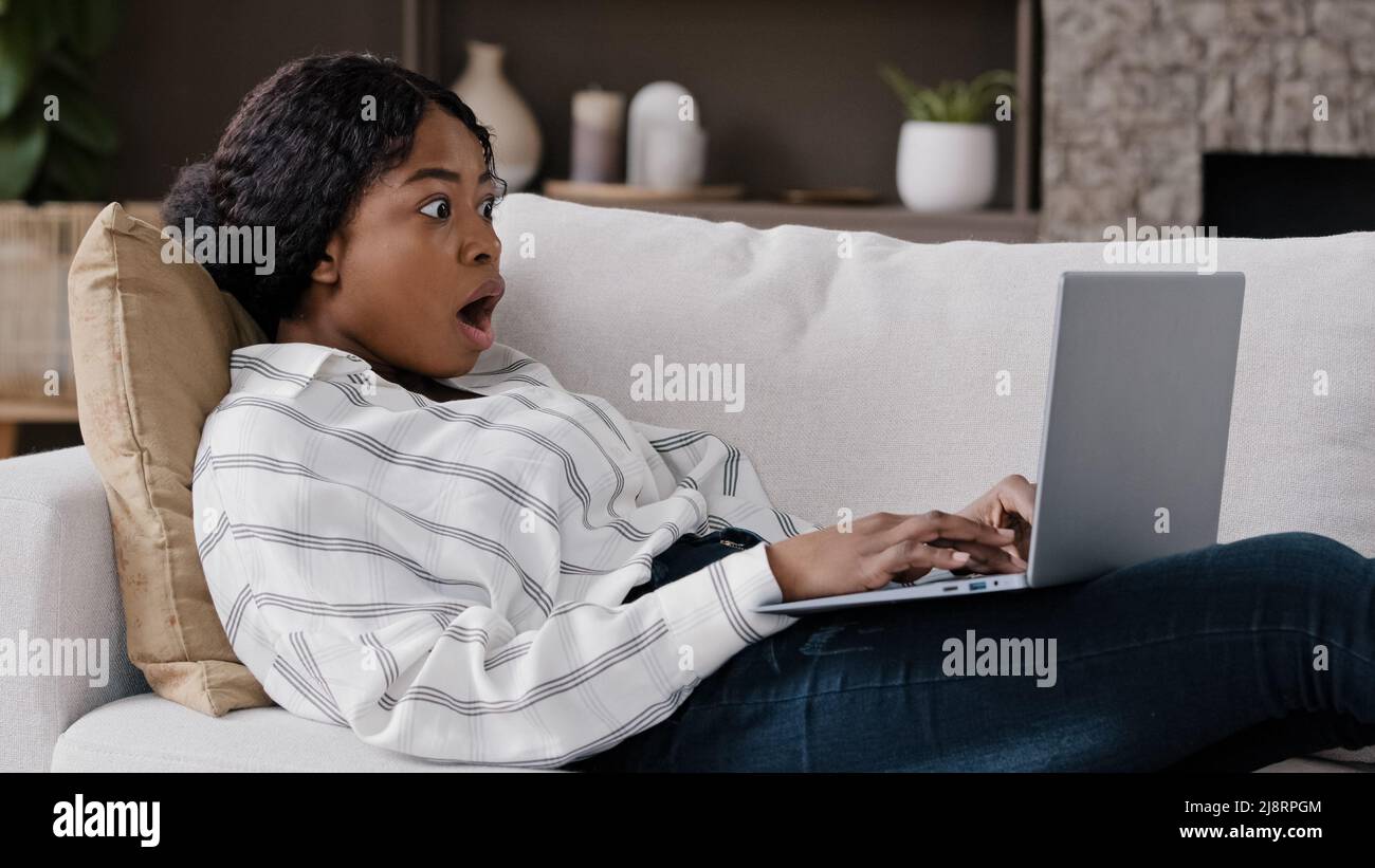 African girl freelancer american biracial woman lying on couch at home looking in laptop reading unexpected bad news gossip receiving notification ope Stock Photo