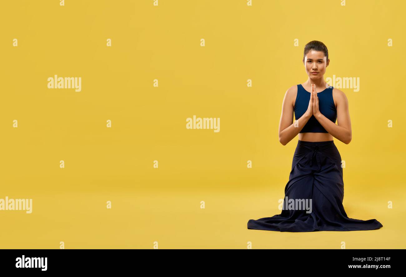 Female yoga instructor sitting on knees in prayer pose in spacious studio. Front view of brunette girl kneeling and praying, looking at camera, isolat Stock Photo