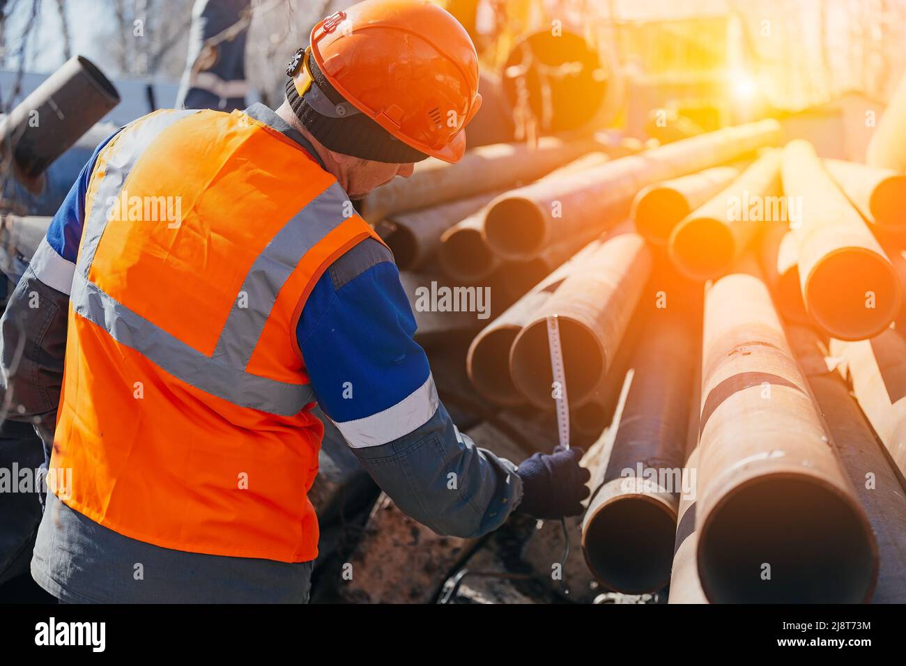 Builder in helmet and vest measures diameter of pipe. Workflow. Open warehouse with metal pipes. sun glare, Stock Photo