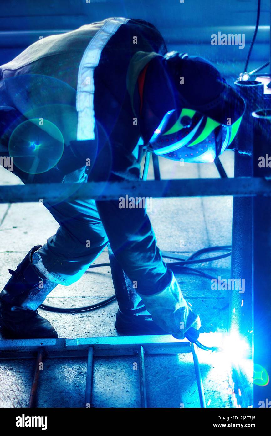 Professional welder in protective uniform and mask works in production hall. Authentic workflow. Welder works with metal and sparks fly. Stock Photo