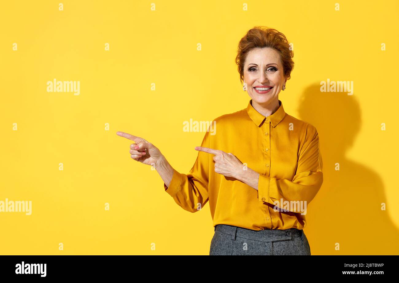 Smiling woman points away with both forefingers, shows on copy space for your text. Photo of attractive elderly woman in yellow shirt on yellow backgr Stock Photo