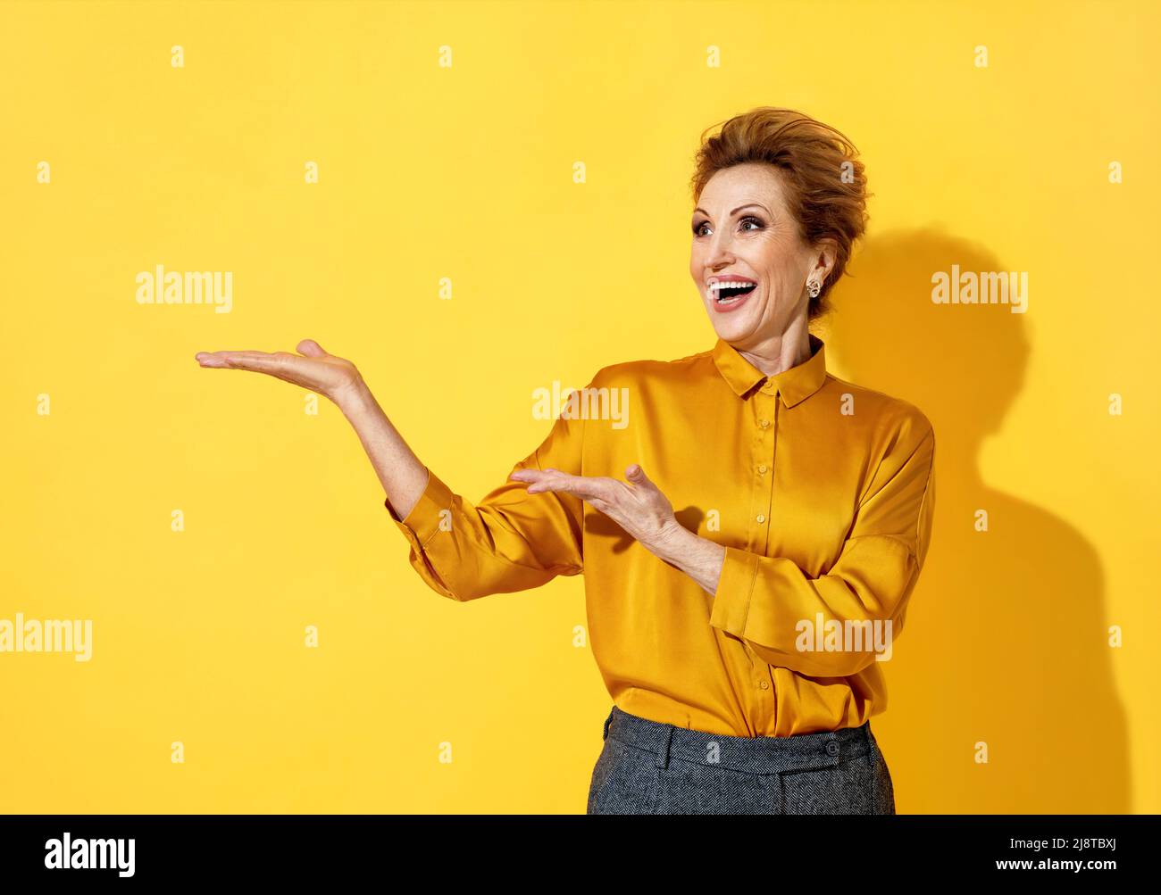 Cheerful woman showing empty copy space (ads, promo, recommend). Photo of attractive elderly woman in yellow shirt on yellow background Stock Photo