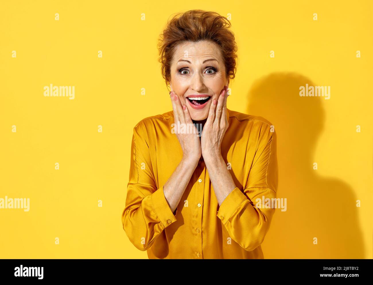 Positive smiling woman with eyes full of happiness, holds hands near her face. Photo of attractive elderly woman in yellow shirt on yellow background. Stock Photo
