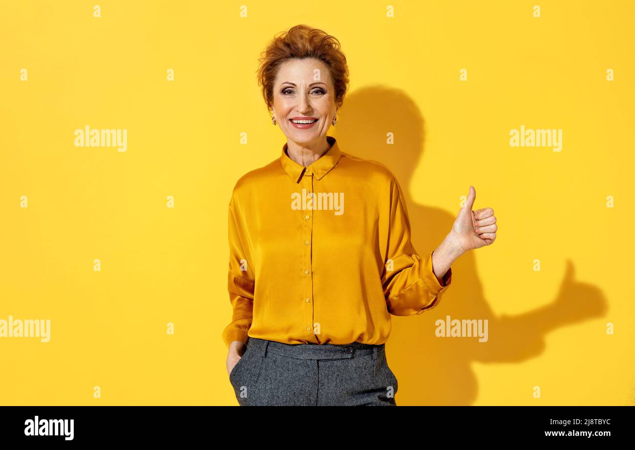 Happy elderly woman shows thumb up, like gesture. Photo of woman in yellow shirt on yellow background. Emotions and pleasant feelings concept. Stock Photo