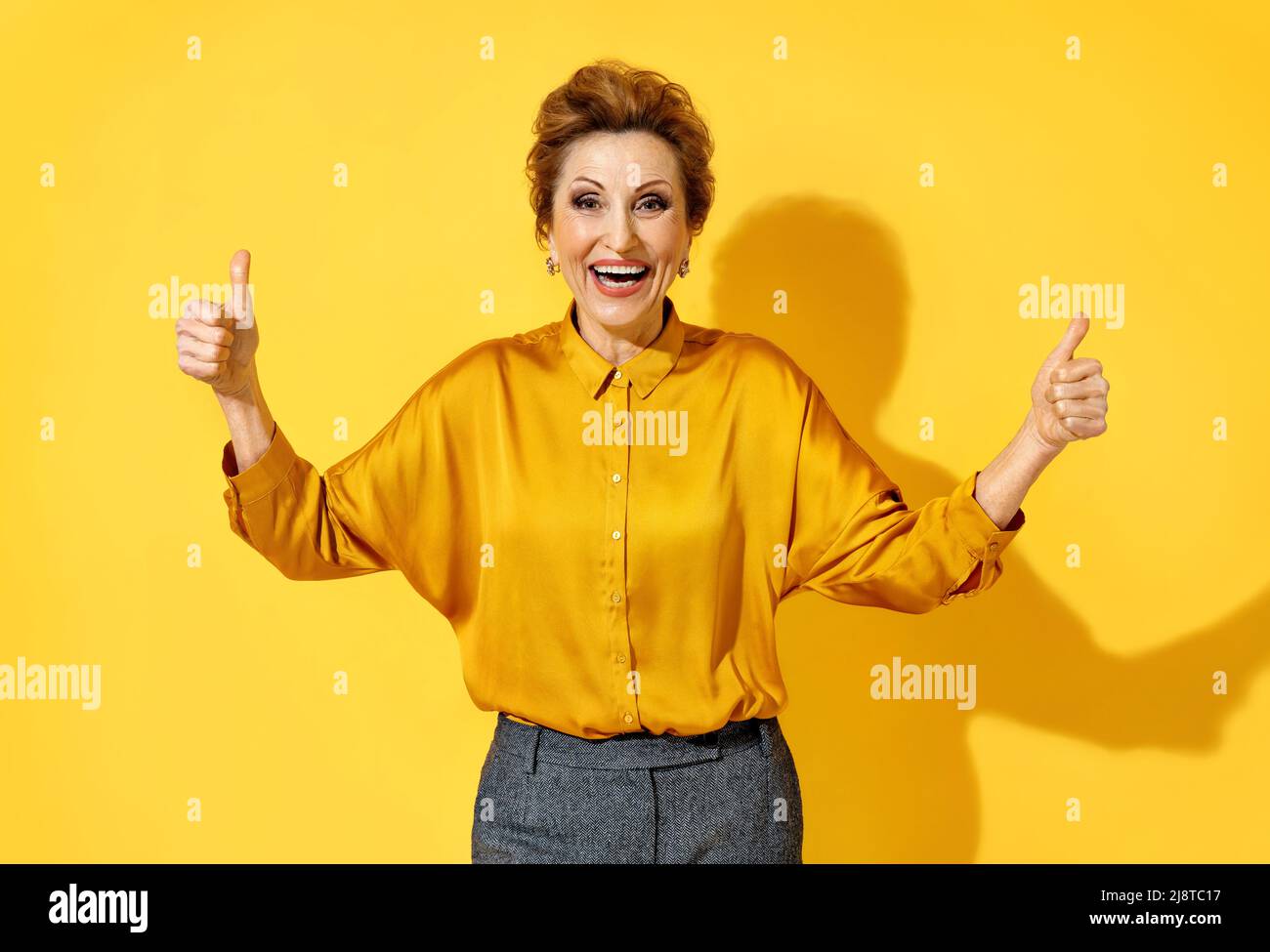 Happy woman shows thumbs up with both hands. Photo of attractive elderly woman in yellow shirt on yellow background Stock Photo