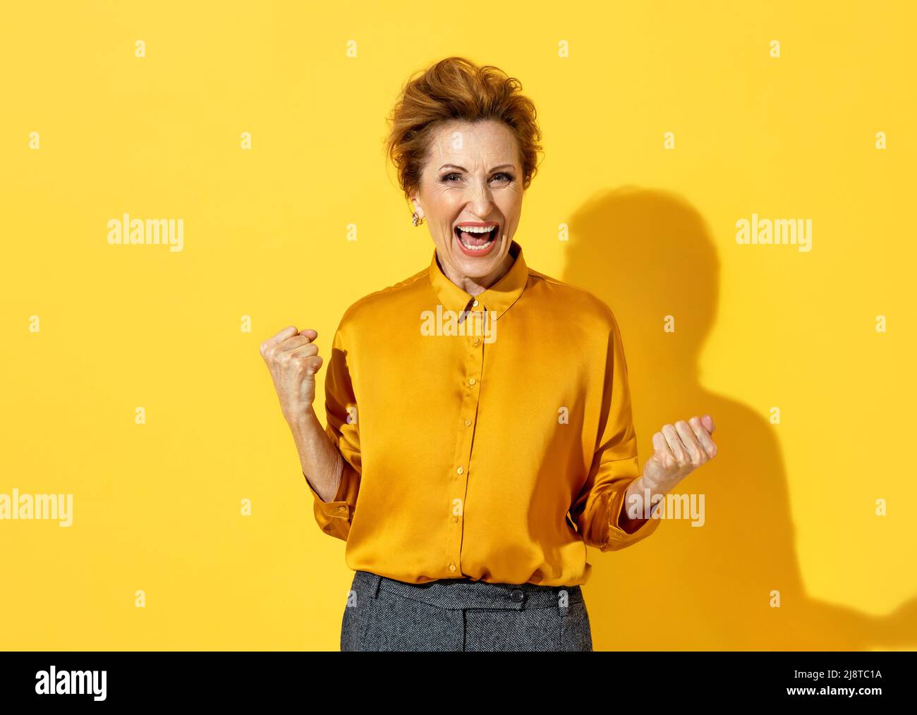 Overjoyed woman clenches fists with happiness, widely opened mouth as shouts loudly. Photo of positive elderly attractive woman in yellow shirt on yel Stock Photo