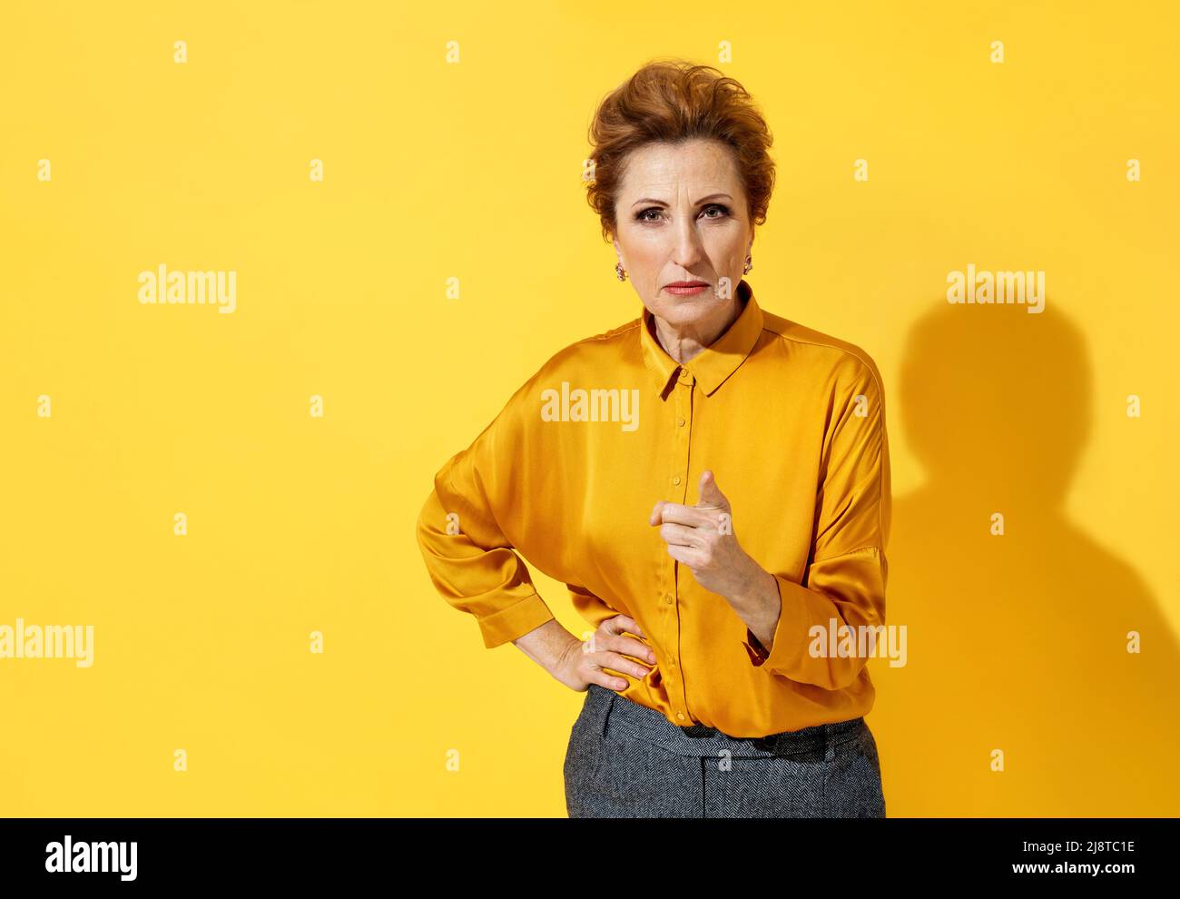 Angry elderly woman scolding someone and points her finger at you, warns, notifies, recommends. Photo of attractive woman in yellow shirt on yellow ba Stock Photo