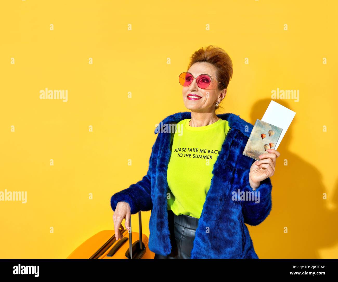 Happy woman is ready to travel. Photo of elderly woman wears pink glasses, dressed in fashionable outfit, with tickets and suitcase on yellow backgrou Stock Photo