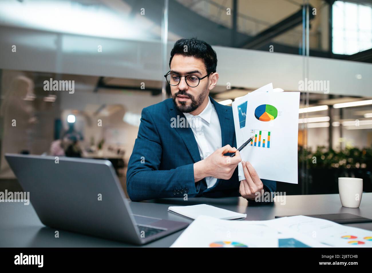Serious busy millennial arab guy with beard in glasses, suit sits at table shows color chart in computer webcam Stock Photo