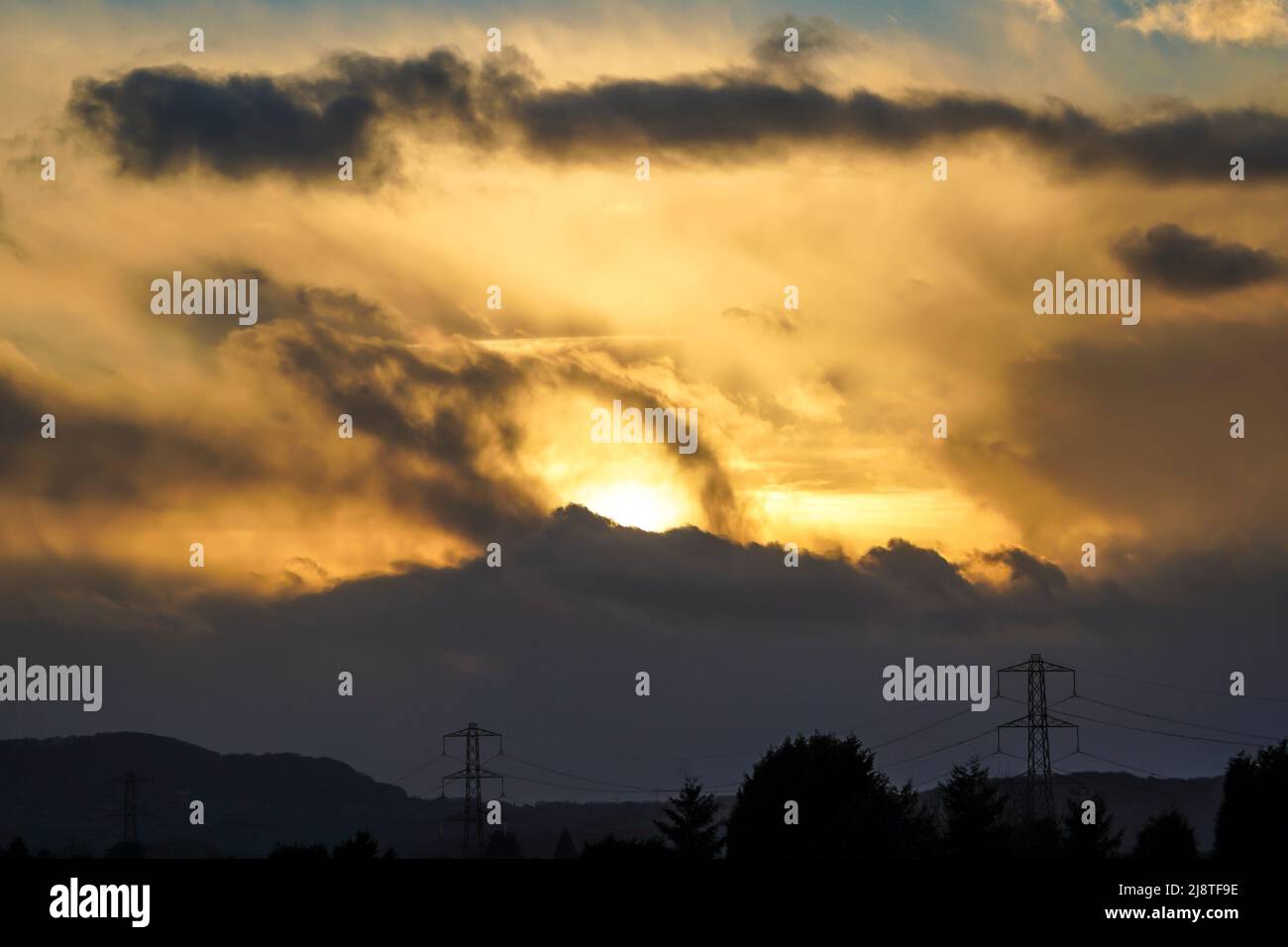 Evening sun setting behind dark clouds over silhouetted electricity pylons, UK. Stock Photo
