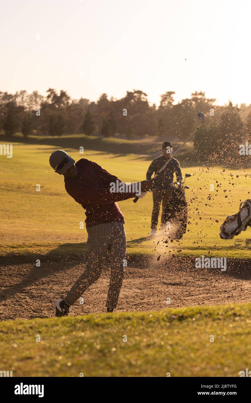 African american young man hitting golf shot while playing with caucasian male friend at course Stock Photo