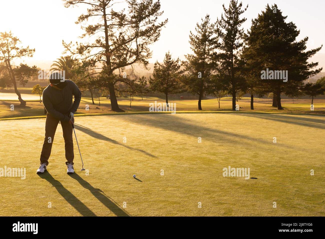 Full length of caucasian young man hitting golf ball with club against trees and clear sky at sunset Stock Photo