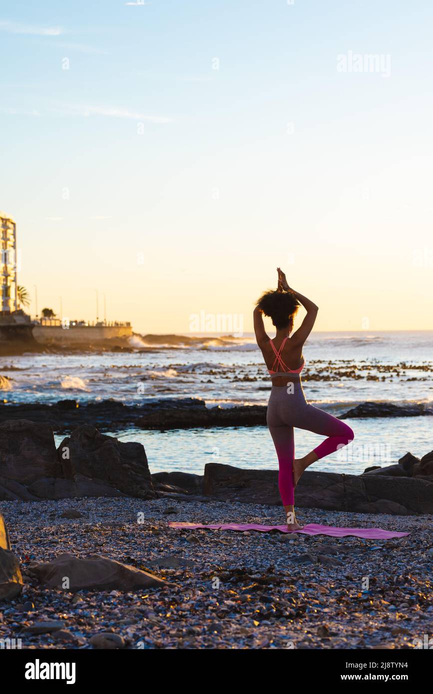 Young african american woman standing on one leg while practicing yoga at beach during sunset Stock Photo