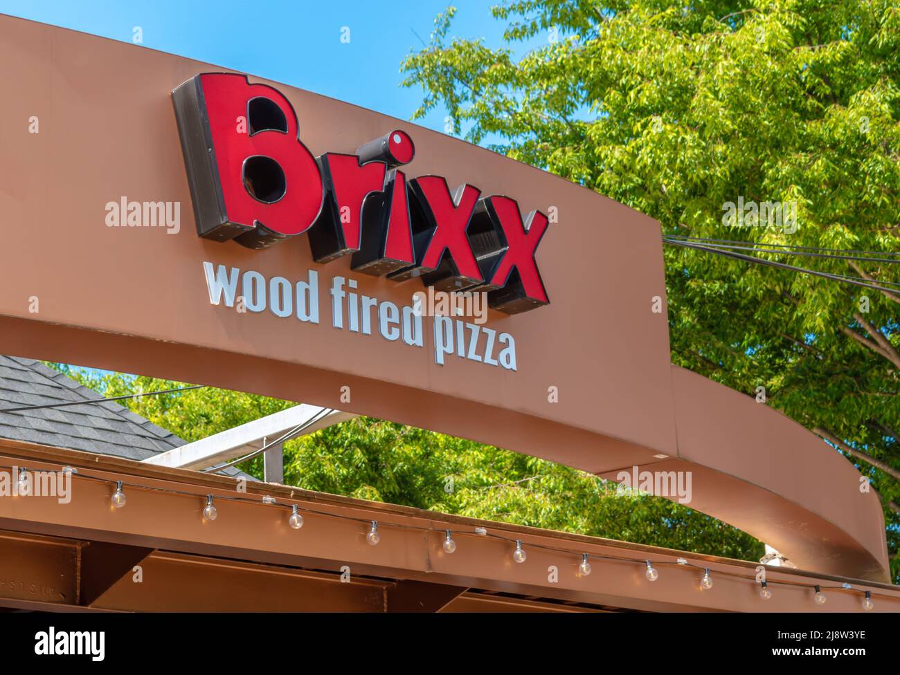 Exterior facade brand and logo signage for 'Brixx' wood fired pizza store on a bright sunny day with shadows, green trees leaves and a clear blue sky. Stock Photo