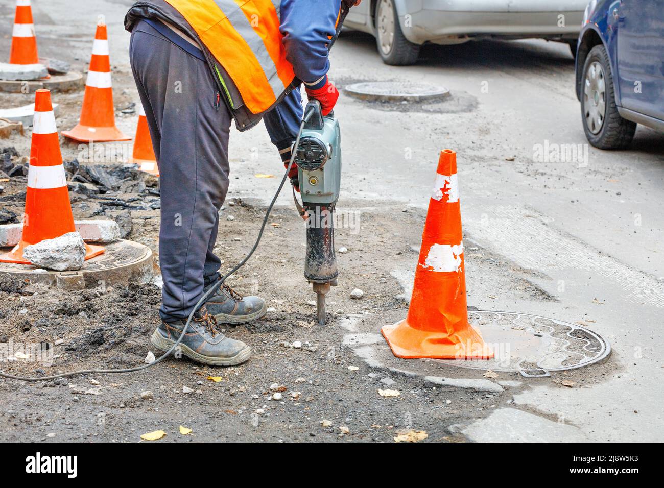 A road worker works with an electric jackhammer on a road section fenced with traffic cones. Stock Photo