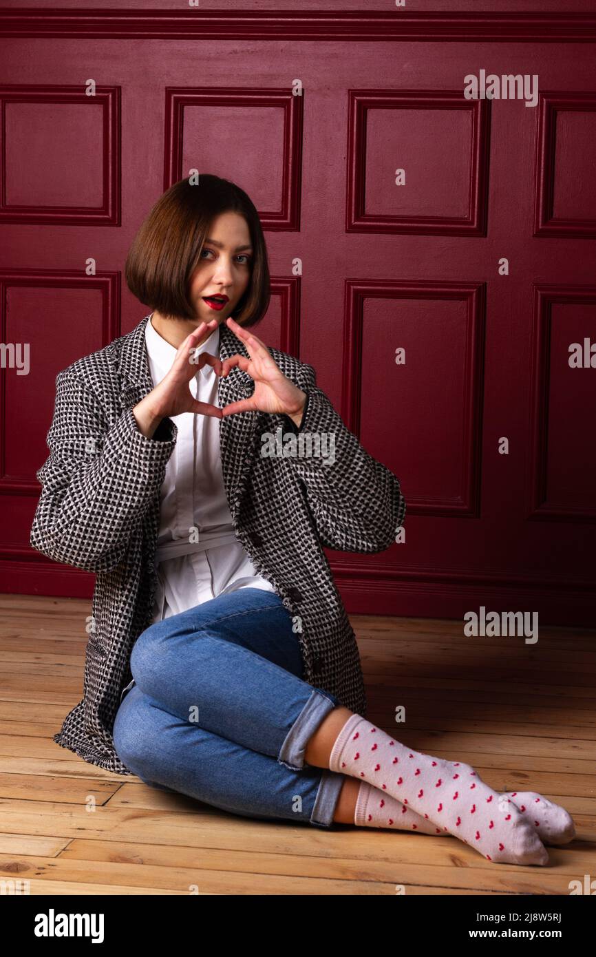 Short-haired woman in white shirt and jacket on red background shaping heart with fingers sitting on floor Stock Photo