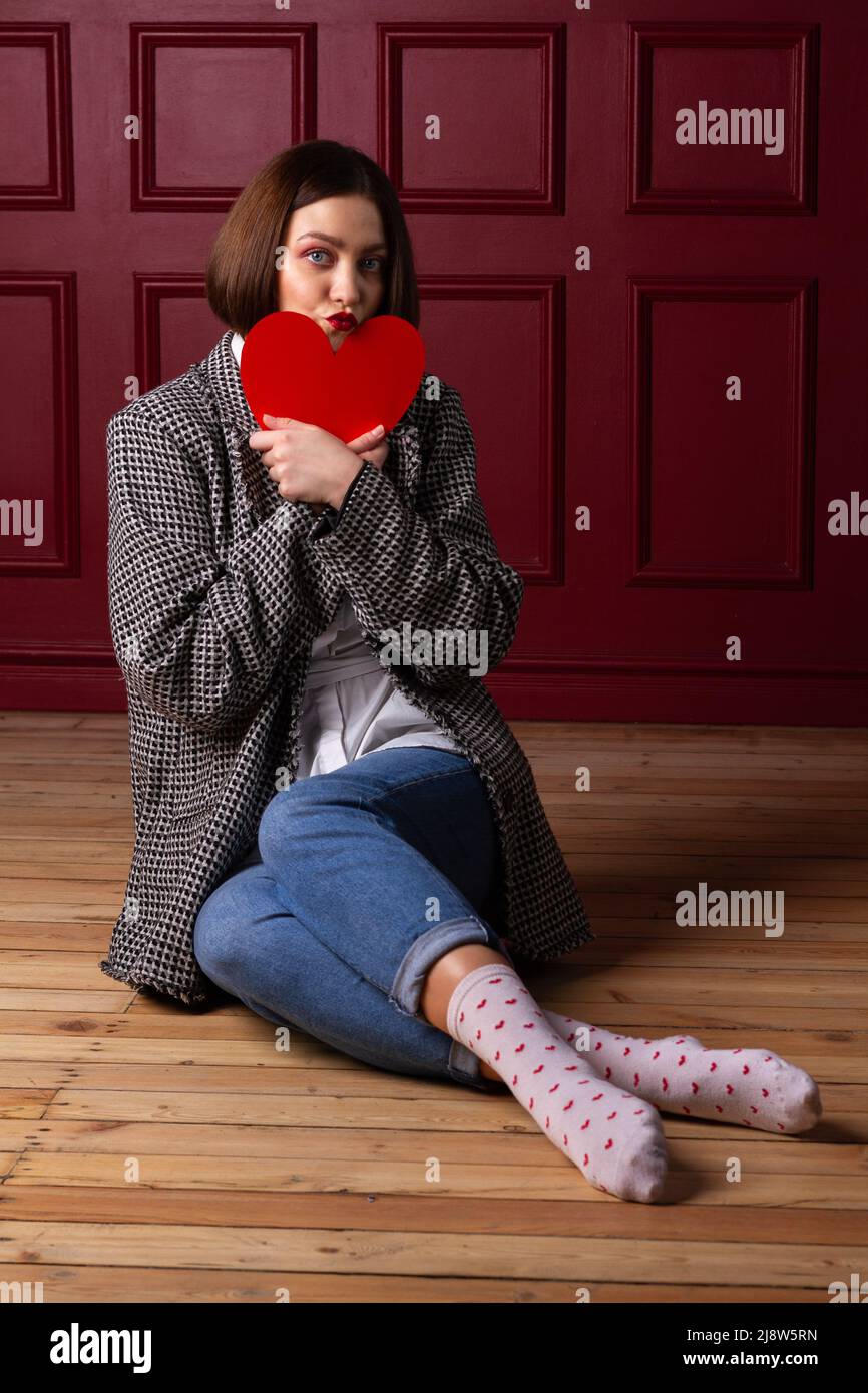 Short-haired woman in jacket sitting on wooden floor and red background holding red heart shape in front of her chin Stock Photo