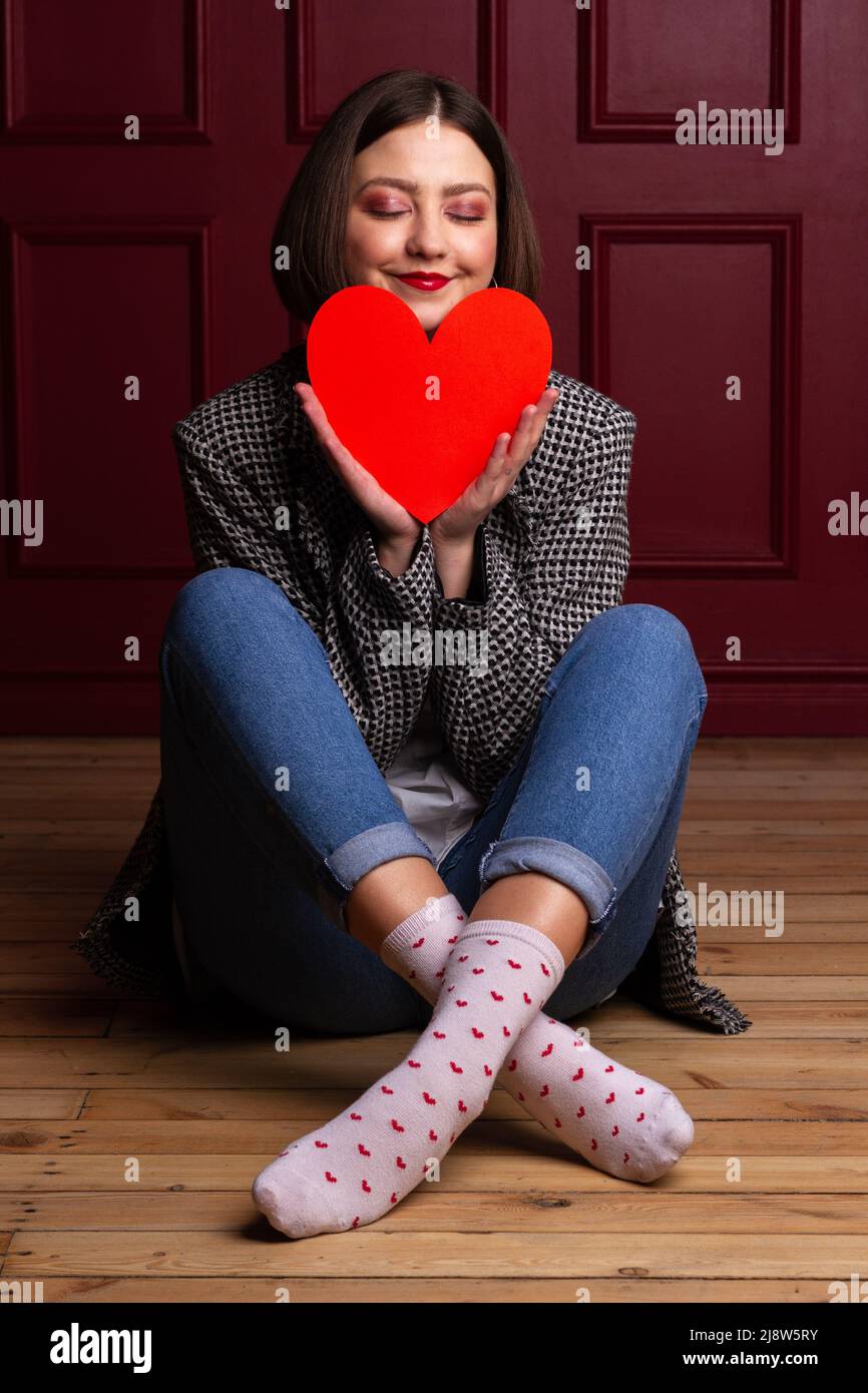 Smiling short-haired woman in jacket sitting on wooden floor legs crossed and red background holding red heart shape in front of her chin Stock Photo