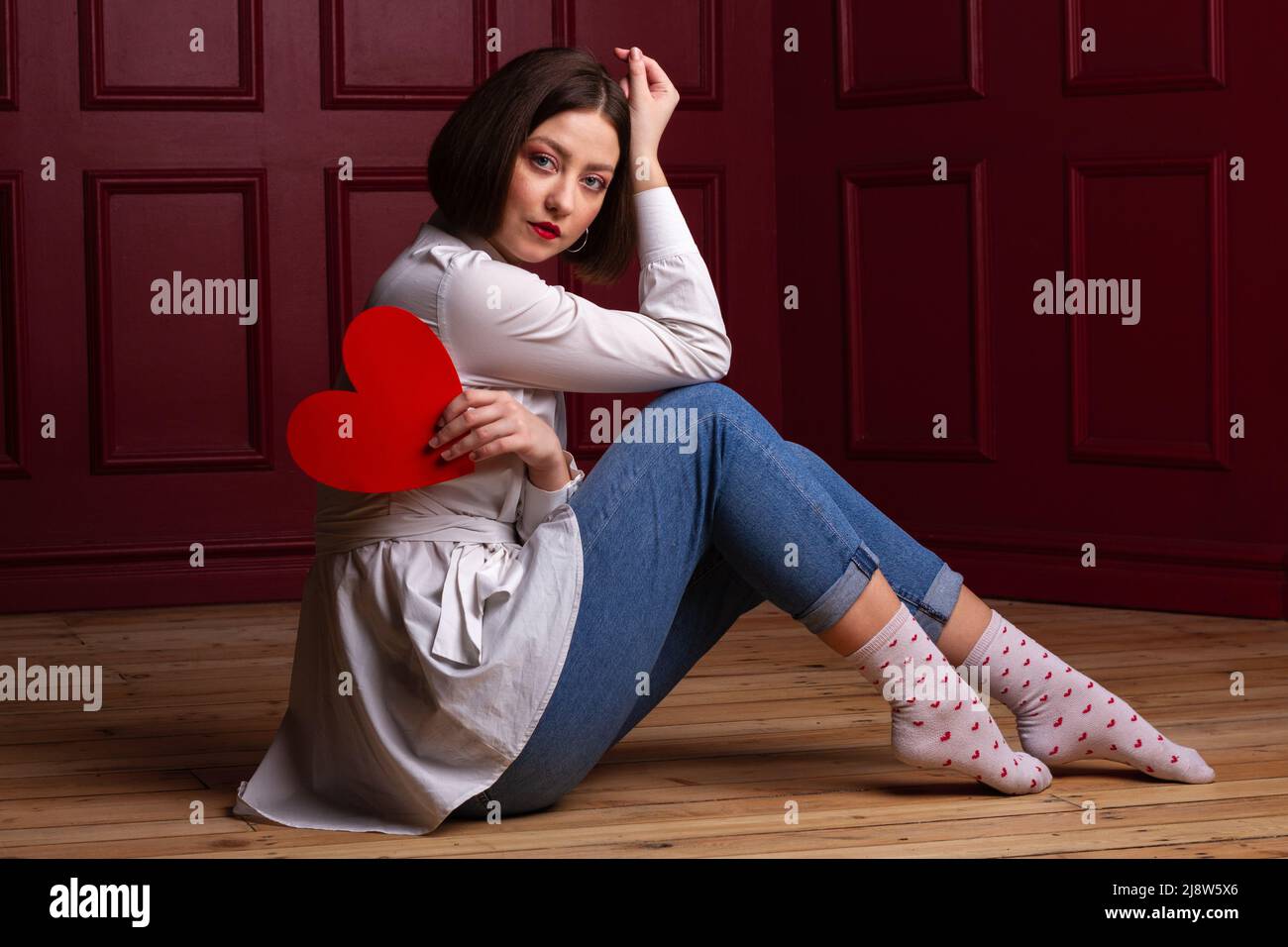 Short-haired woman with head resting on hand sitting side to a camera on a wooden floor and red background holding a red heart shape Stock Photo