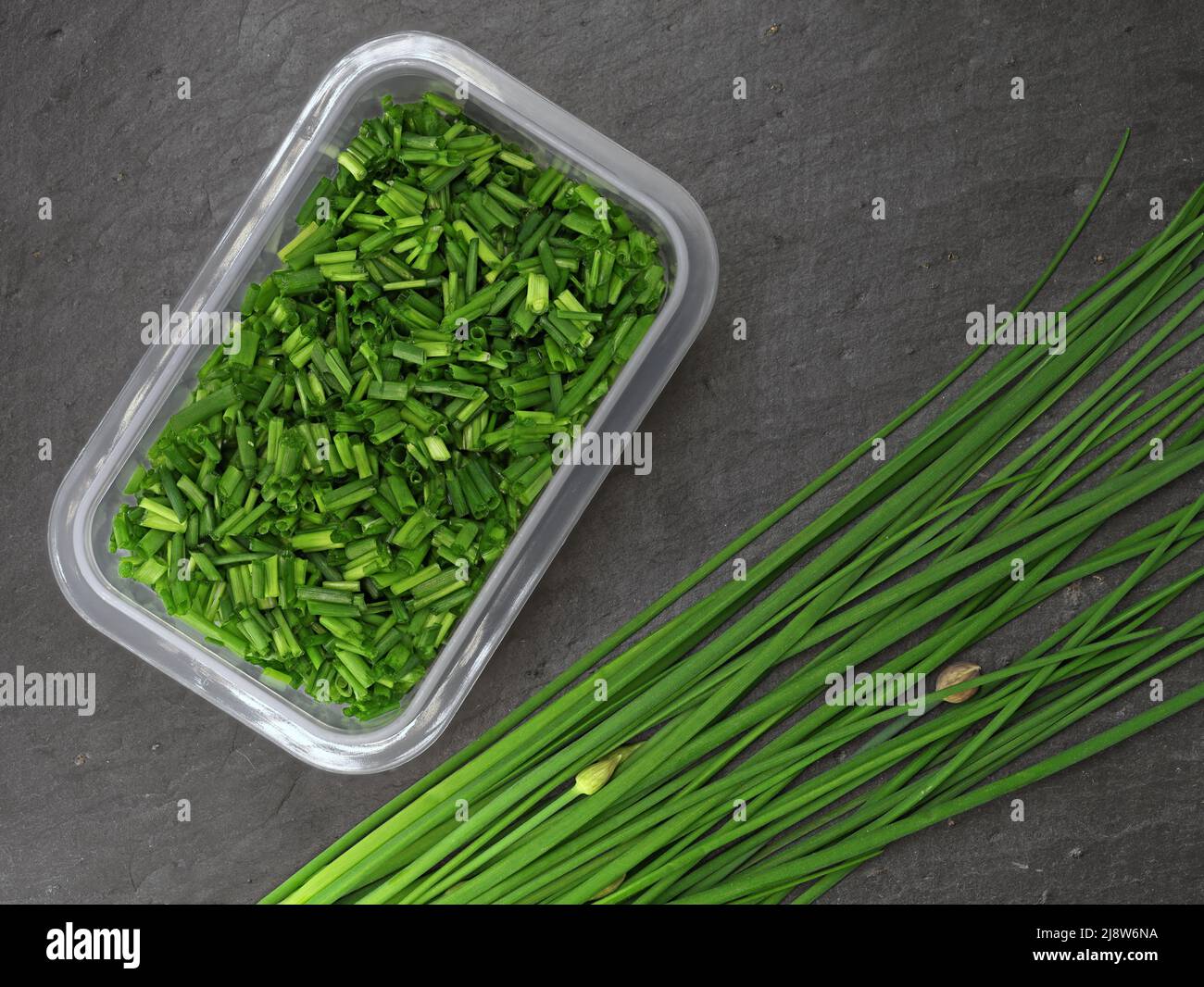 freshly cut chives in storage box next to whole bunch chives on black slate board, top view of