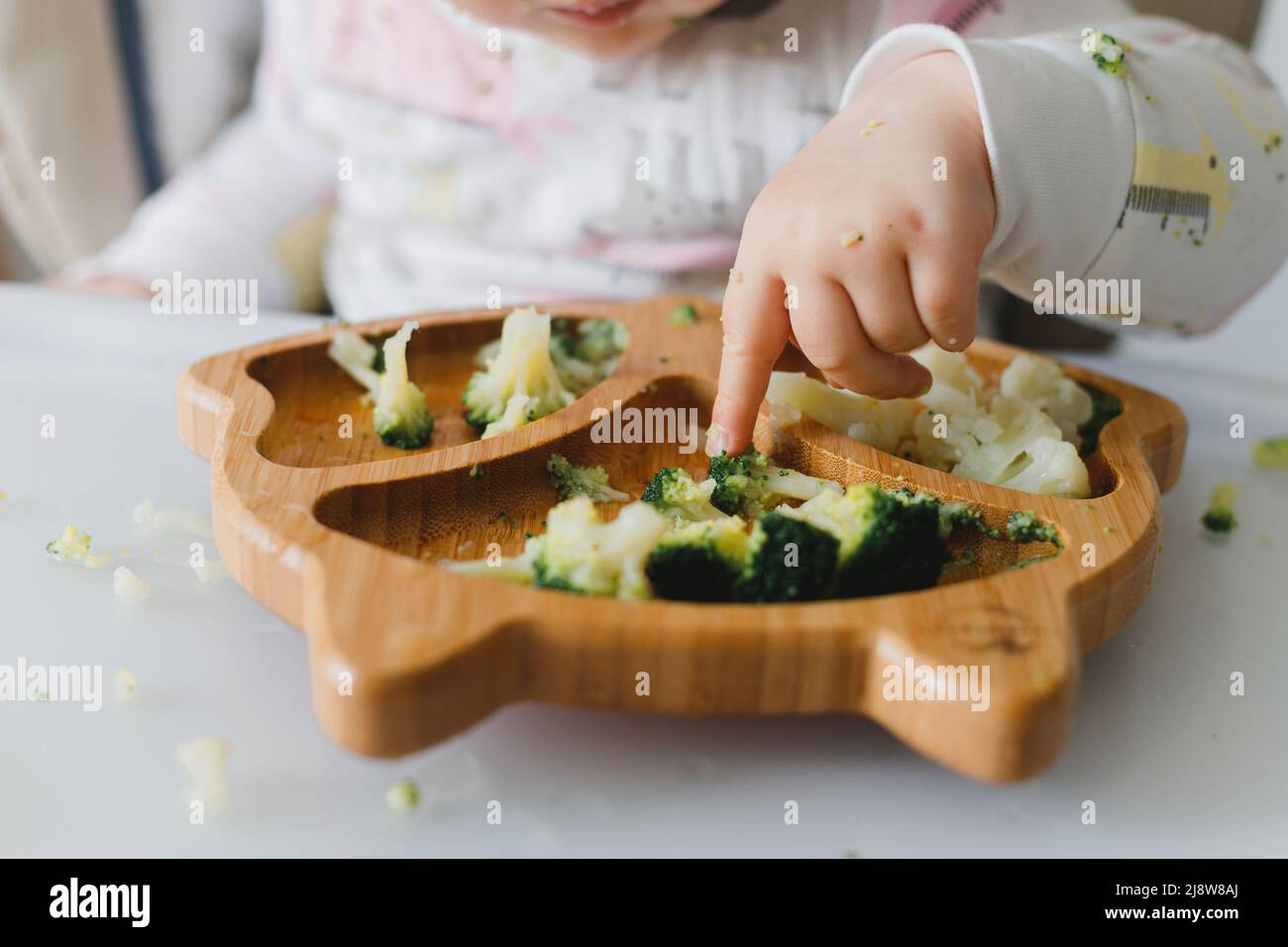 Soiled toddler eats boiled vegetables with his hands awkwardly while sitting on feeding chair Stock Photo