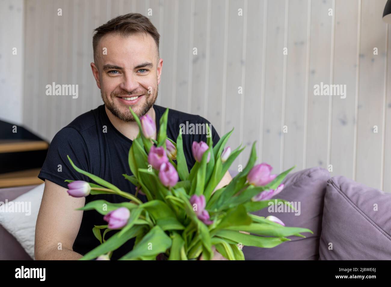 Nice young smiling man in black T-shirt is sitting at home on sofa with bouquet of tulips for his beloved, he looks into camera. Family life Stock Photo