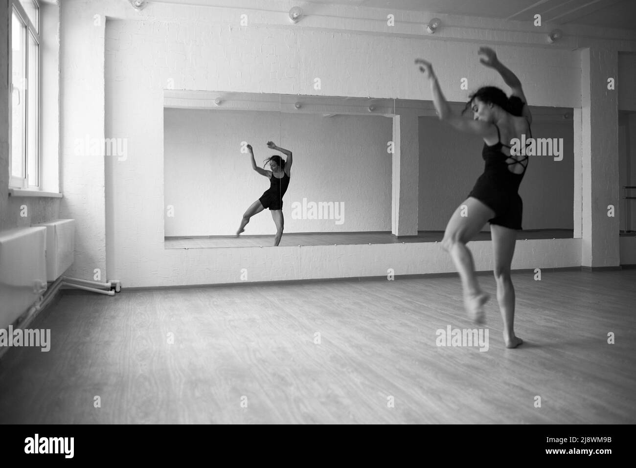 Black and white shot of young female rehearsing modern dance in front of mirror in dancing hall
