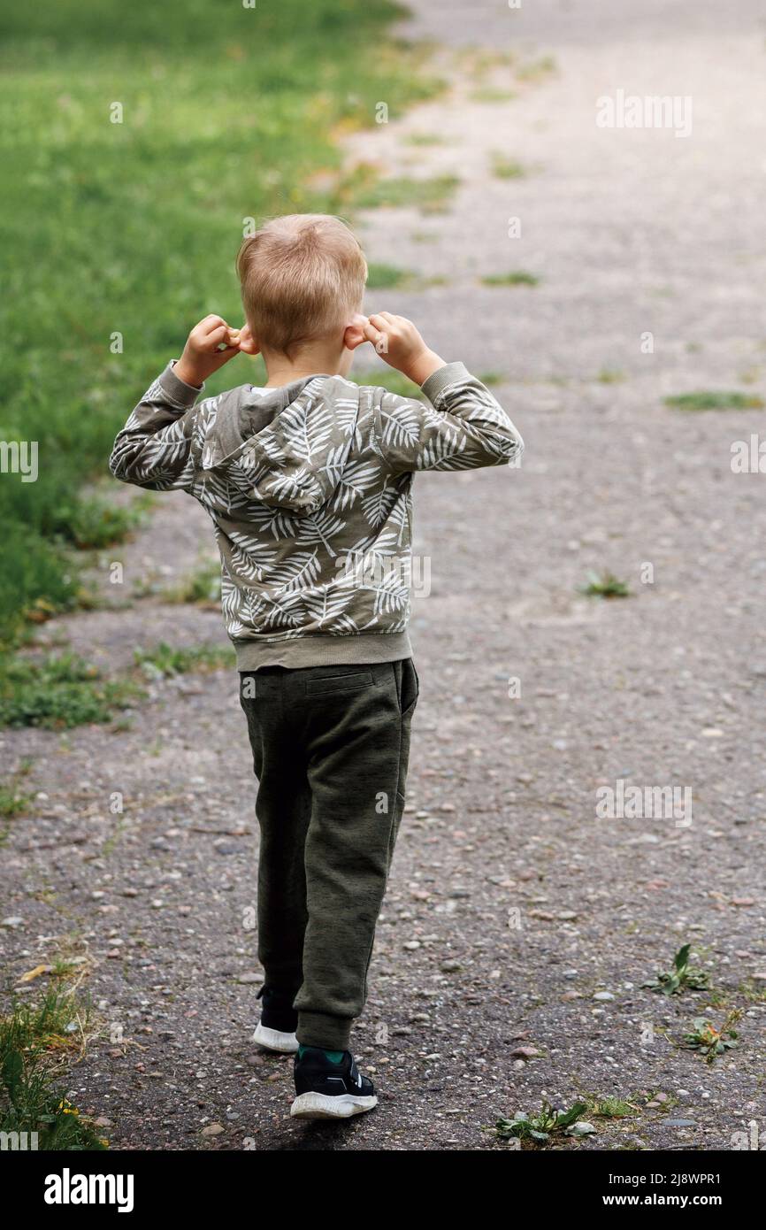 The little boy walks down the road, (rear view) he stretches his ears hard, the child experiments with his body. Stock Photo