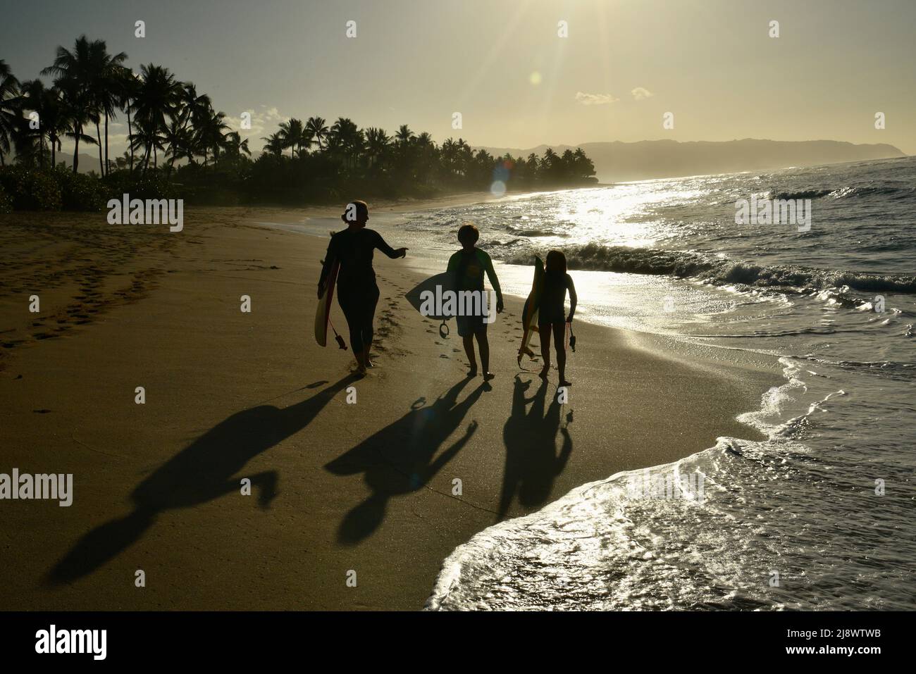 Silhouette of mother and two children walking along beach after surfing during sunset, Haleiwa, Hawaii, USA Stock Photo